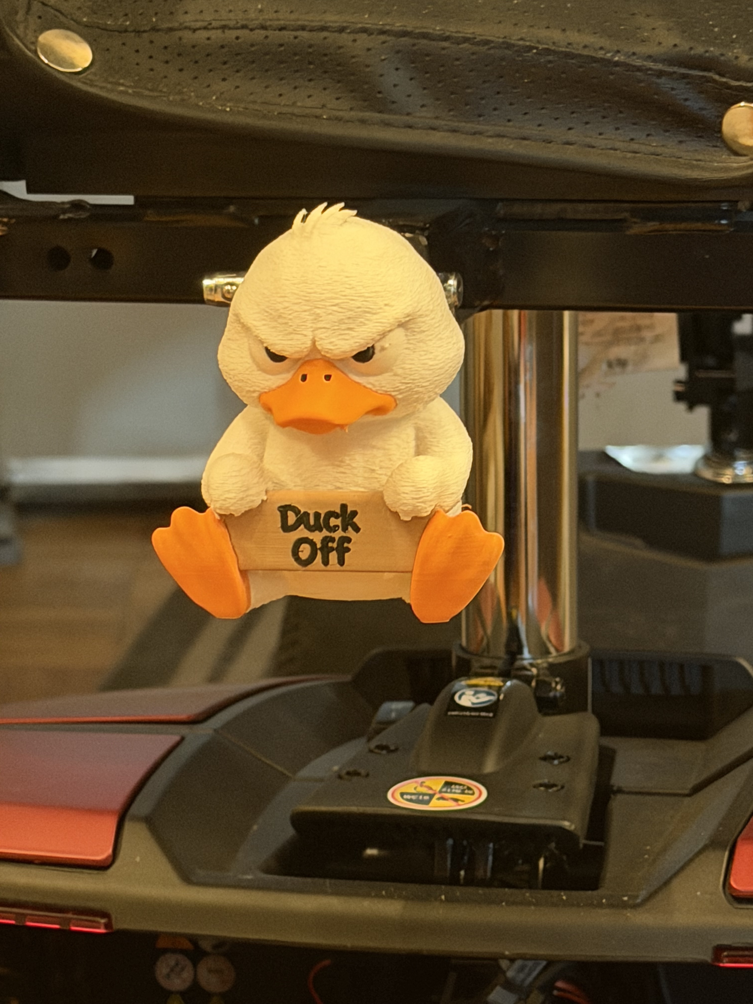 A grumpy plush duck ornament holding a wooden "Duck Off" sign, mounted under the seat of a mobility scooter